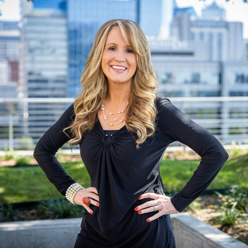 Headshot of Anna Cody, standing with her hands on her hips and wearing a black long-sleeved dress, with a city skyline in the background