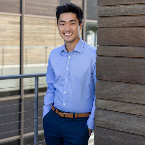 Headshot of Chester Wang, wearing a light blue, patterned, button-up shirt, leaning against a wooden building