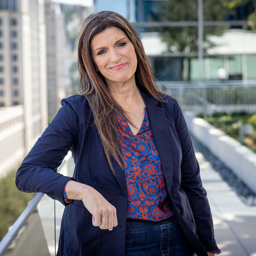 Headshot of Debbie McBride, wearing a solid, dark blue jacket over a floral blouse, with a city skyline in the background
