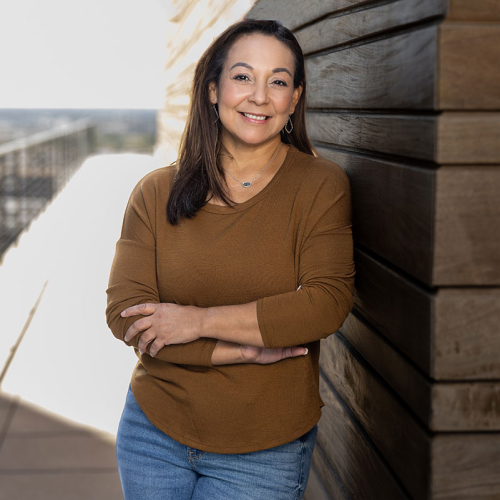 Headshot of Irma Guzman, wearing a tan long-sleeved shirt, leaning against a wooden building