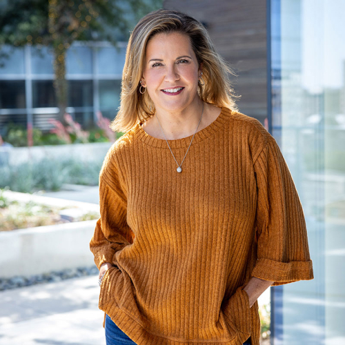 Headshot of Laura Beagle, wearing an orange sweater, with a business park courtyard in the background