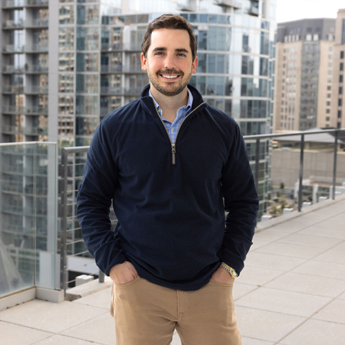 Headshot of Matthew Tramonte, wearing a solid, dark blue, zip-up sweater, with a city skyline in the background