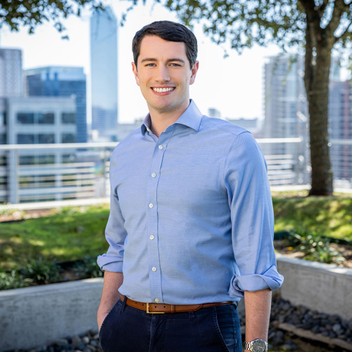 Headshot of Travis Monteleone, wearing a light blue, button-up shirt with the sleeves rolled up, with a city skyline in the background