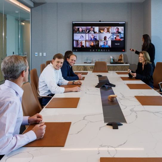Group of six NGP employees dressed in business attire sitting at a marble conference table and working together with their fellow employees on a video conference call.