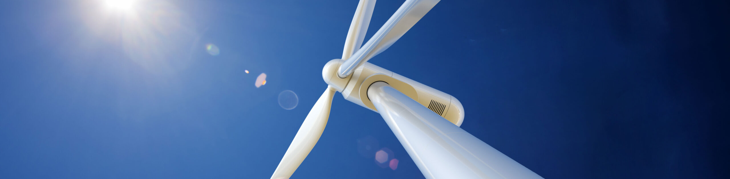 Close-up shot of a white wind turbine in the foreground with a navy blue sky in the background with snippet of the sun shining in on the wind turbine